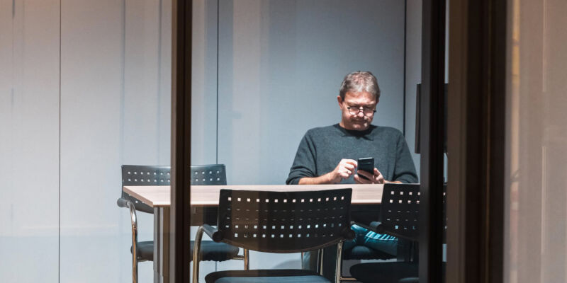 Person sitting alone in a glass meeting room, focused on his phone or work